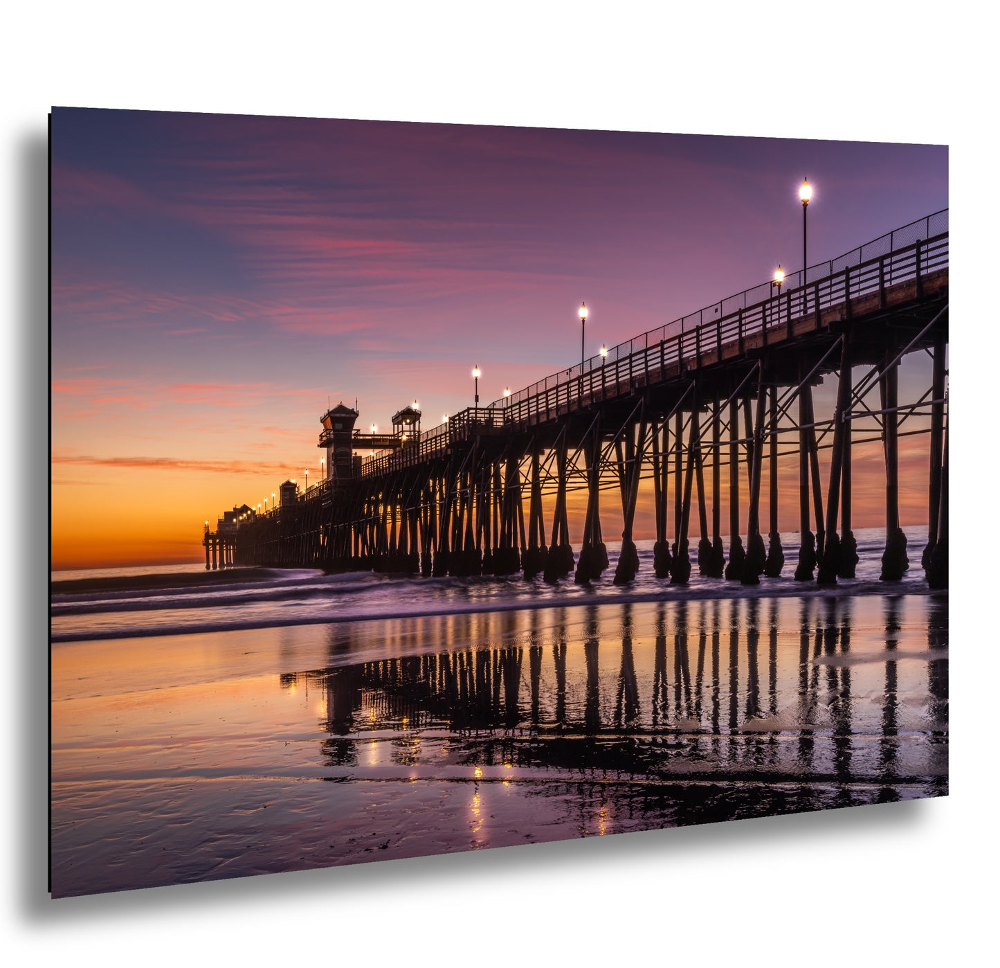 A picturesque pier extending into the ocean at sunset, with its reflection mirrored in the calm waters. The sky is painted in vibrant shades of orange and pink, creating a stunning backdrop for the scene. The pier is illuminated by warm, glowing lights, adding to the overall ambiance of the image.