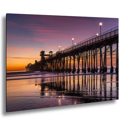 A picturesque pier extending into the ocean at sunset, with its reflection mirrored in the calm waters. The sky is painted in vibrant shades of orange and pink, creating a stunning backdrop for the scene. The pier is illuminated by warm, glowing lights, adding to the overall ambiance of the image.