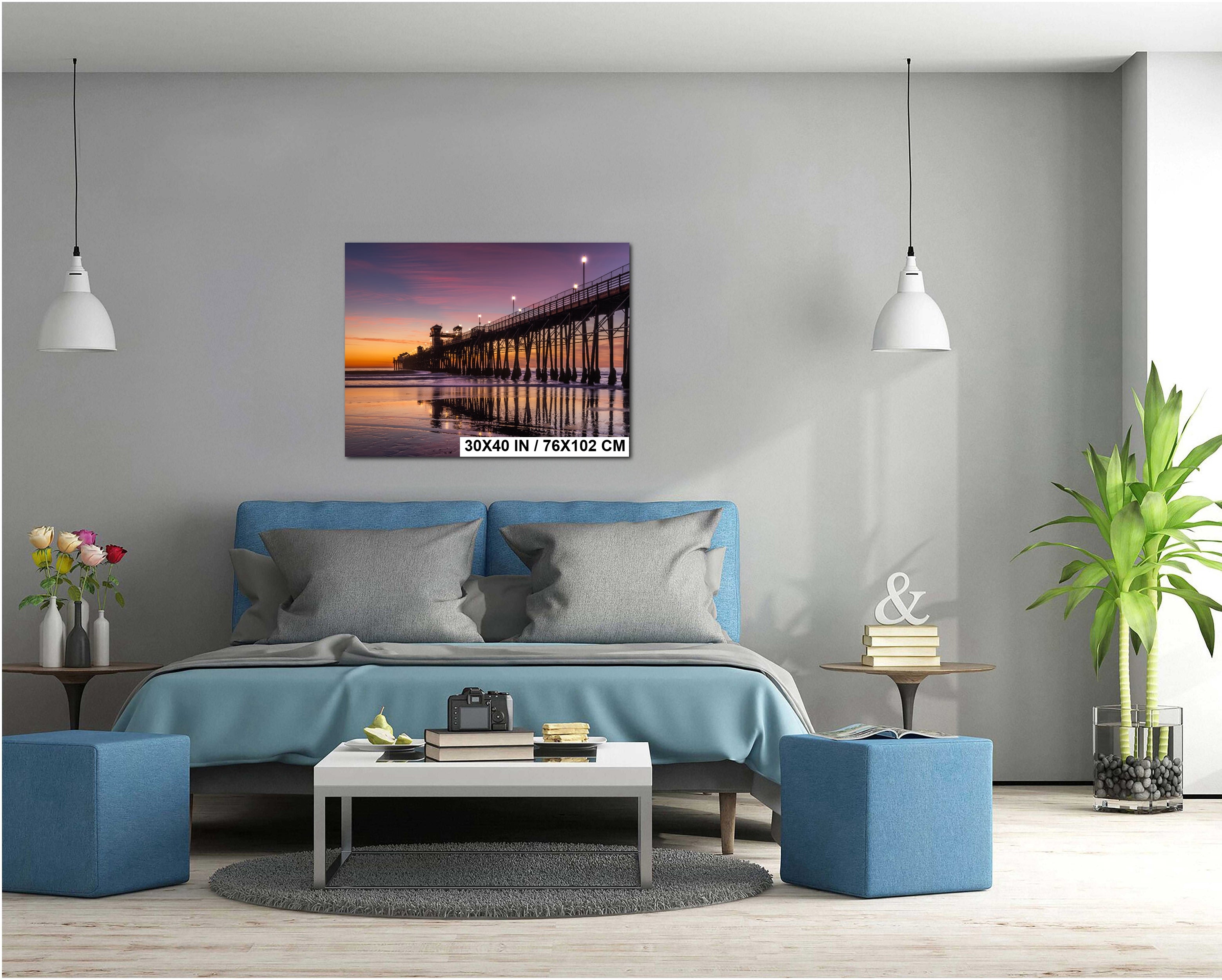 a serene bedroom scene with a large bed, a nightstand, and a coffee table. The bed is covered in a blue comforter and has two gray pillows. Above the bed, there is a framed photograph of a pier at sunset. The room is decorated with potted plants, vases, and books, creating a cozy and inviting atmosphere.