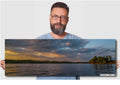 Man holding a panoramic photo of a lake with trees and clouds.