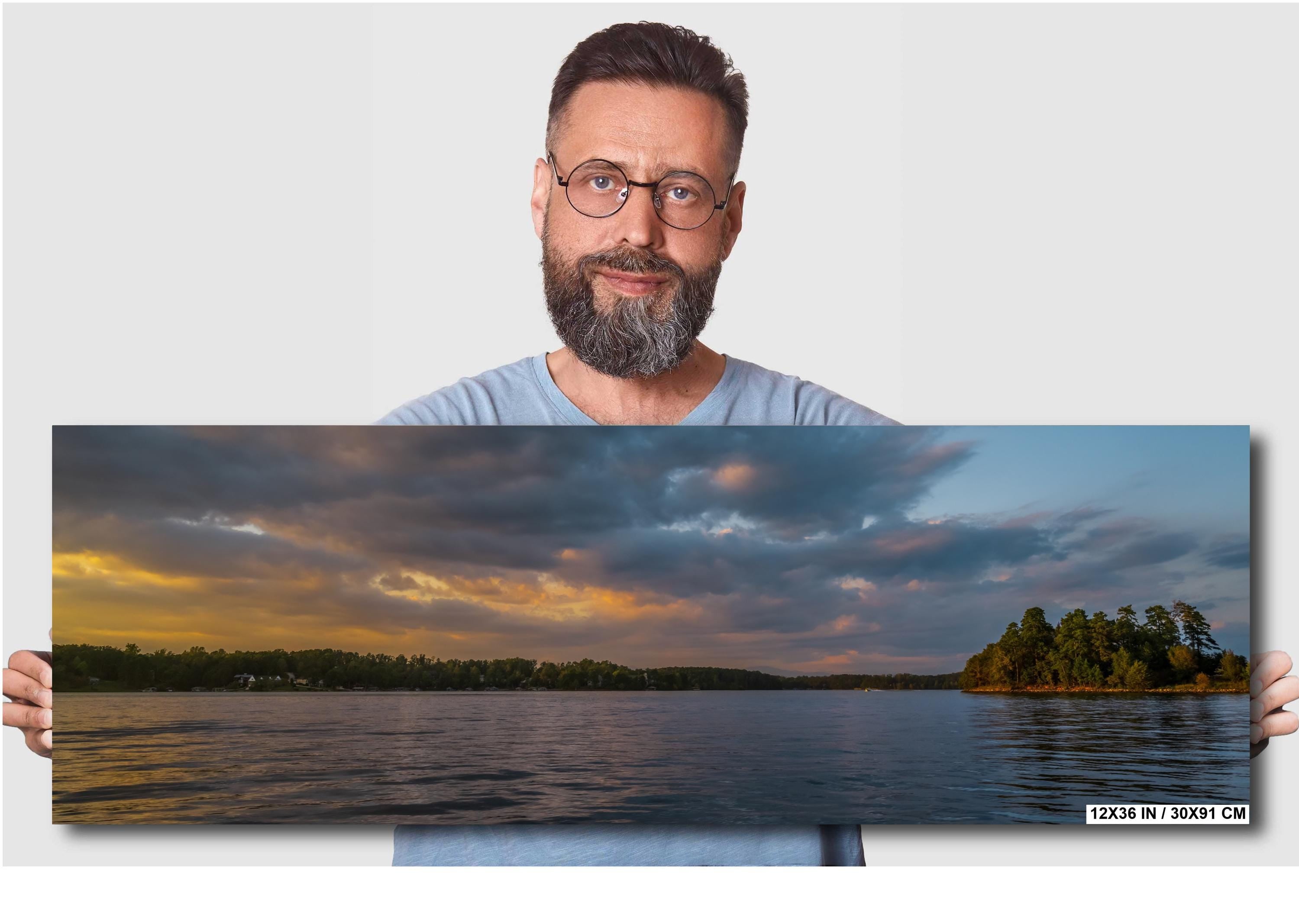 Man holding a panoramic photo of a lake with trees and clouds.