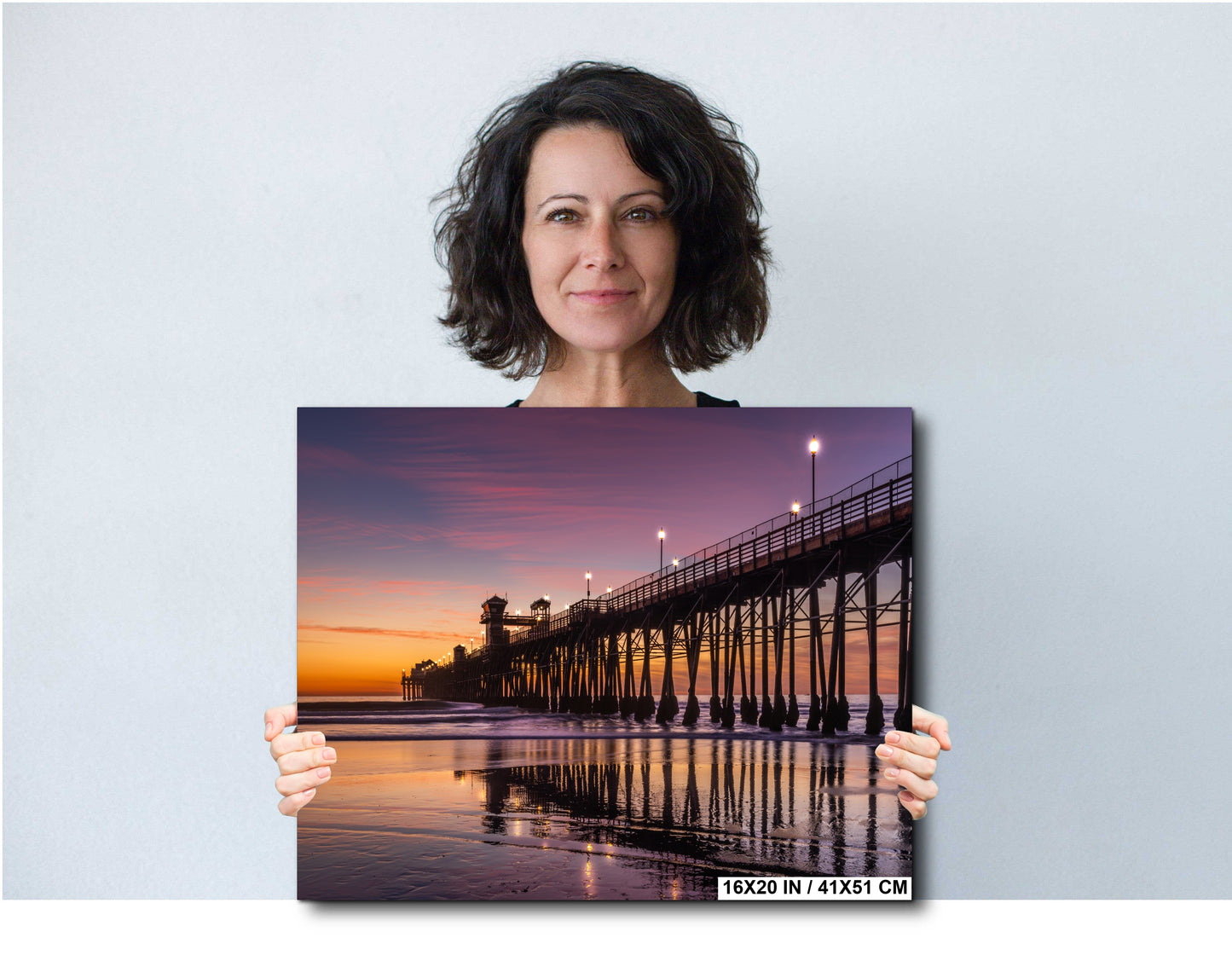 A woman holds a framed print of a serene beach scene at sunset, featuring a wooden pier extending into the water.