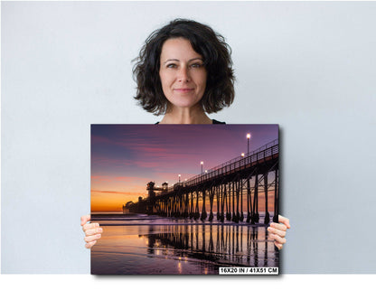 A woman holds a framed print of a serene beach scene at sunset, featuring a wooden pier extending into the water.