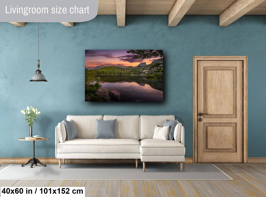 a living room with a large wall painting of a serene landscape featuring a lake and mountains. The painting is hung above a white sofa, which is positioned in front of a wooden door. The room has a wooden floor and a small table with a vase of flowers. The wall painting is approximately 40 inches wide and 15 inches tall, and it is mounted at a height of approximately 101/152 inches.