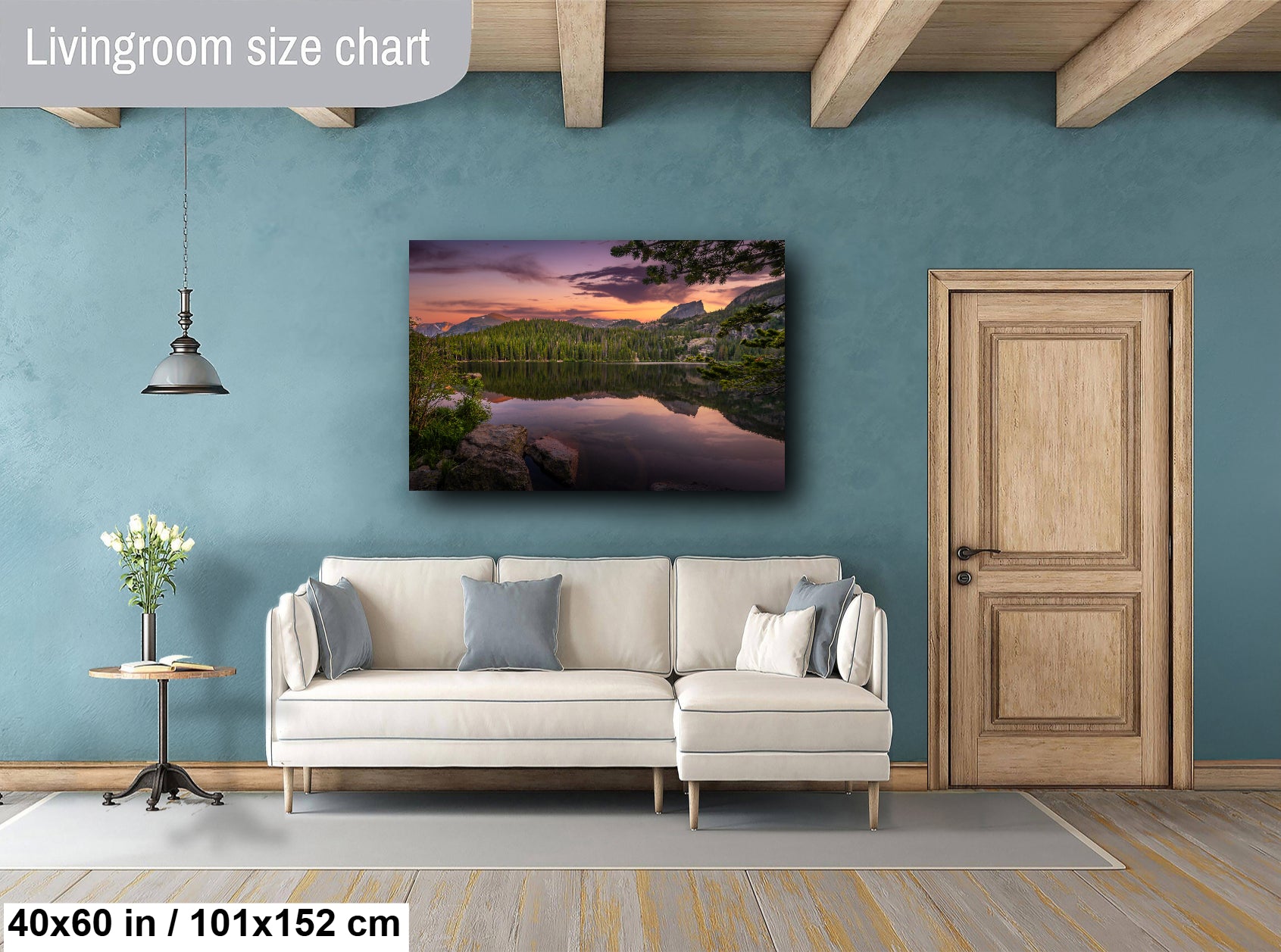 a living room with a large wall painting of a serene landscape featuring a lake and mountains. The painting is hung above a white sofa, which is positioned in front of a wooden door. The room has a wooden floor and a small table with a vase of flowers. The wall painting is approximately 40 inches wide and 15 inches tall, and it is mounted at a height of approximately 101/152 inches.
