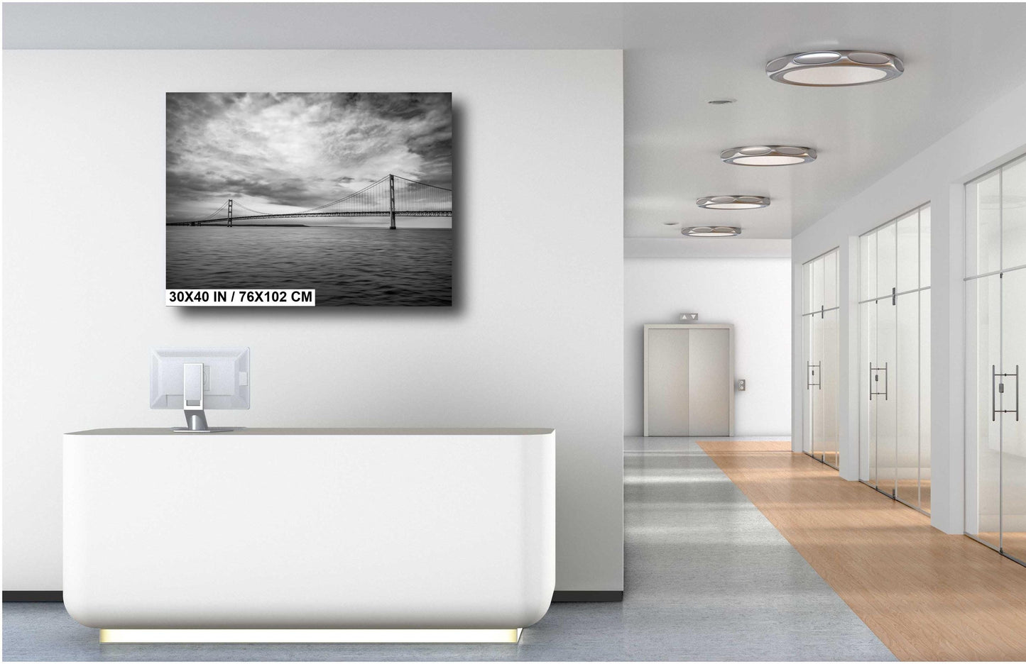 A 30x40 inch black and white horizontal art print of a suspension bridge over water, mounted on a white wall above a modern white reception desk in a bright, minimalist office hallway.