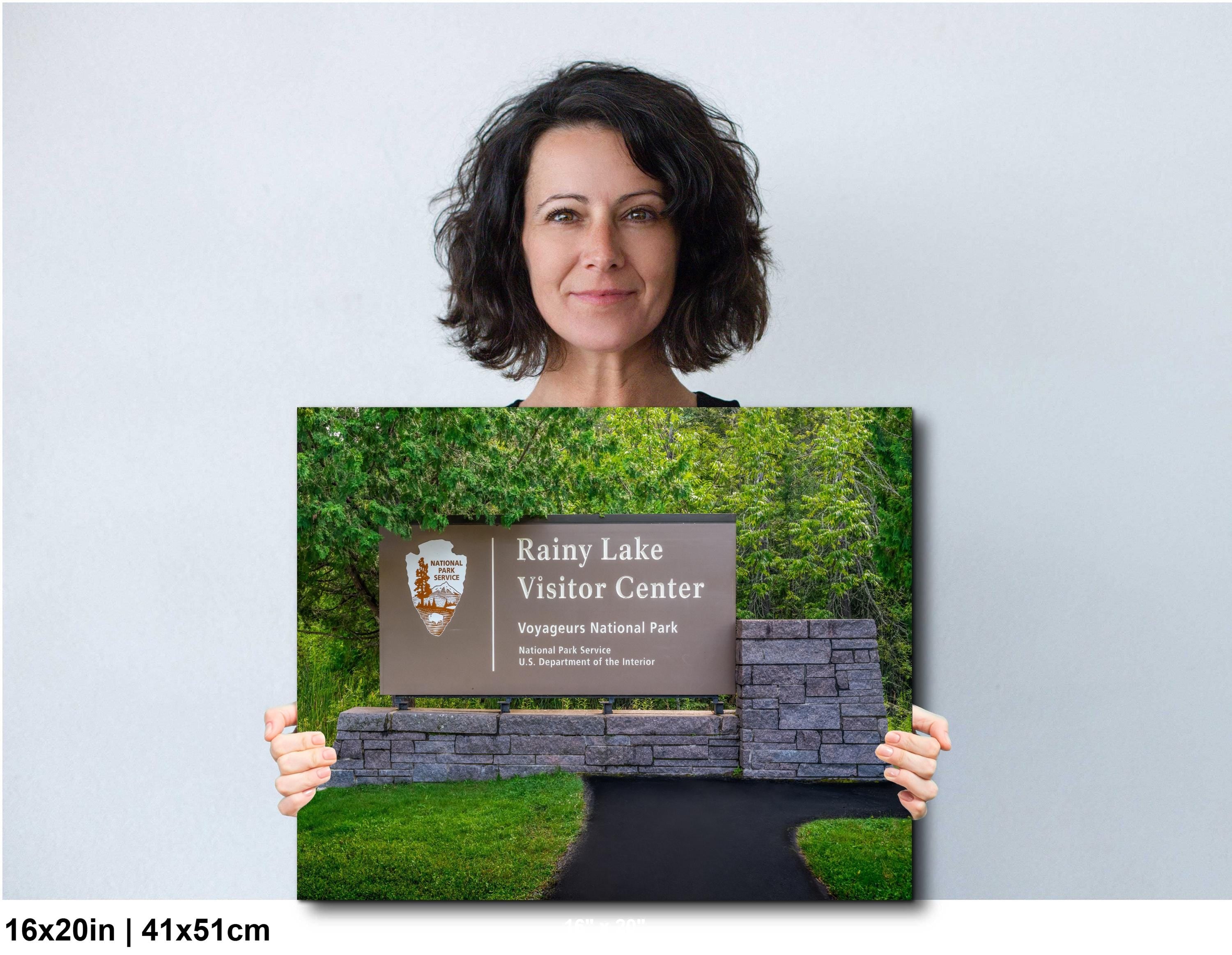 Woman holding a print of Rainy Lake Visitor Center sign at Voyageurs National Park with stone base and trees.