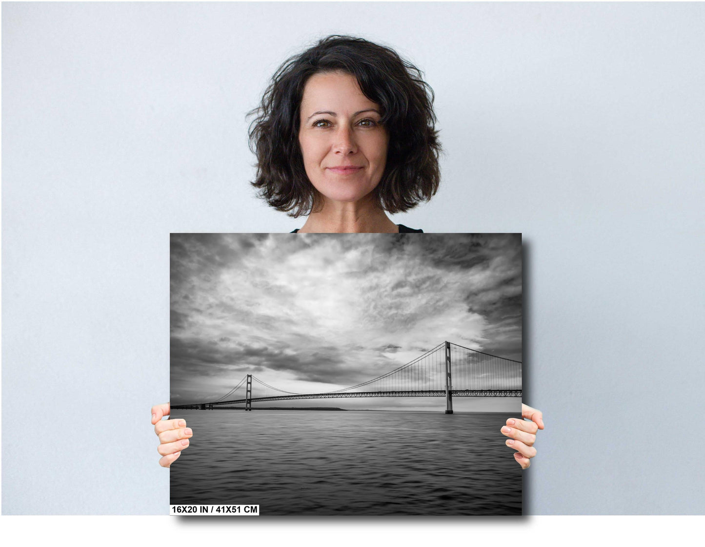 A woman holding a 16x20 inch (41 x 51 cm) black and white landscape photograph of a suspension bridge over water against a light grey background.