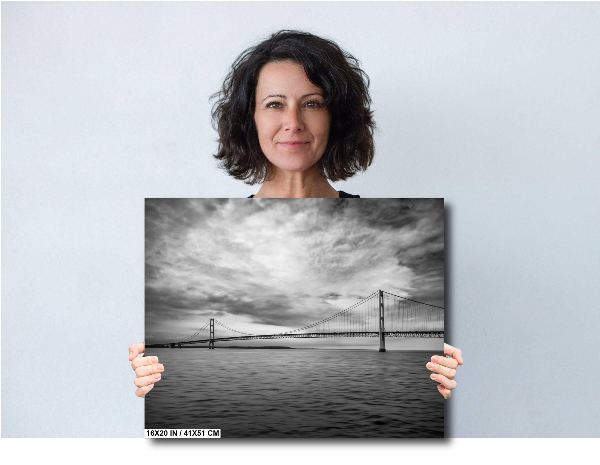 A woman holding a 16x20 inch (41 x 51 cm) black and white landscape photograph of a suspension bridge over water against a light grey background.