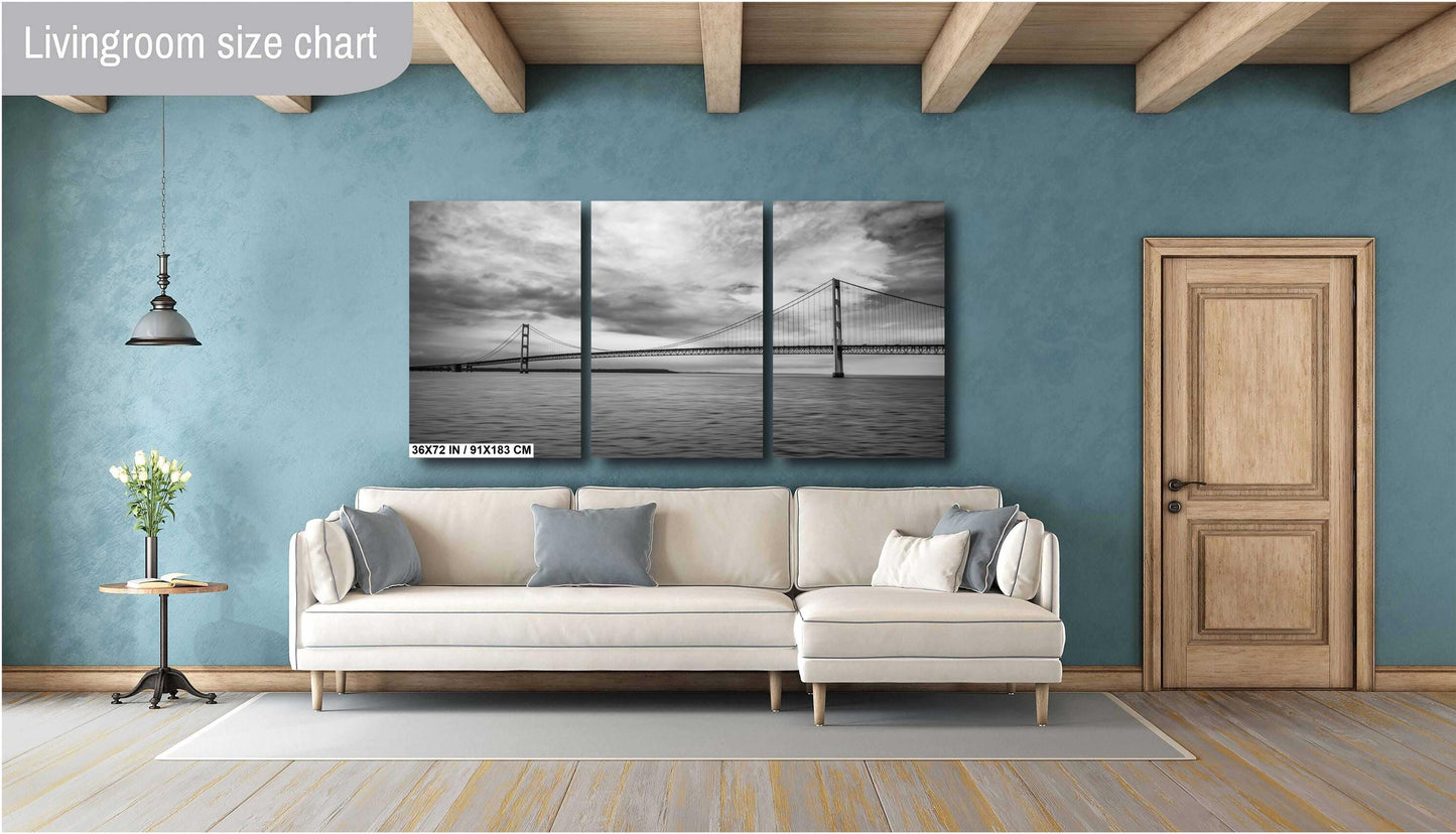 A three-panel vertical triptych art print of a black and white suspension bridge over water, measuring 36x72 inches, mounted on a teal blue wall above a modern cream-colored sectional sofa in a stylish living room with wooden ceiling beams.