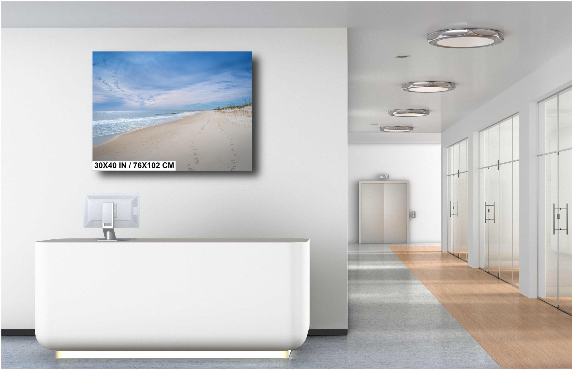 A 30x40 inch landscape photograph of a serene beach with white sand and blue skies, displayed on a white wall above a modern white reception desk in a bright, minimalist office hallway.