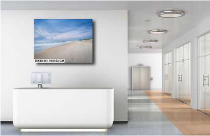 A 30x40 inch landscape photograph of a serene beach with white sand and blue skies, displayed on a white wall above a modern white reception desk in a bright, minimalist office hallway.