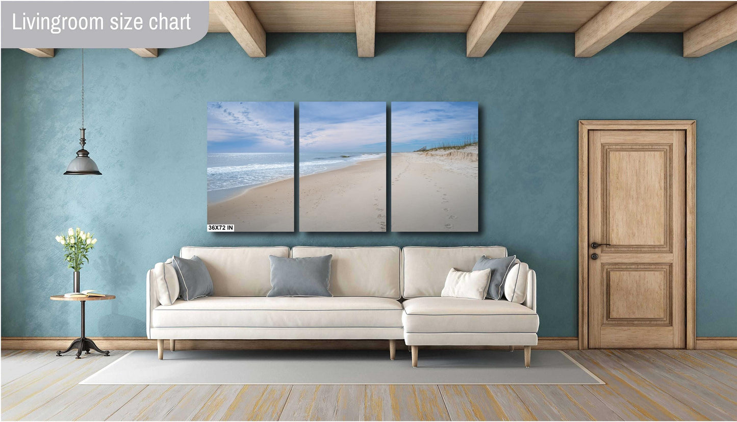 A three-panel triptych landscape photograph of a serene beach at 36x72 inches, mounted on a teal blue wall above a modern cream-colored sectional sofa in a stylish living room with wooden ceiling beams.