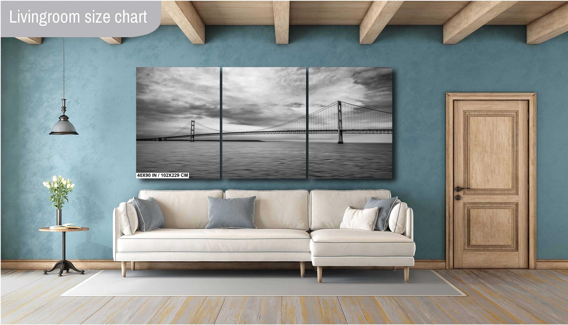 A three-panel vertical triptych art print of a black and white suspension bridge over water, measuring 40x90 inches, mounted on a teal blue wall above a modern cream-colored sectional sofa in a stylish living room with wooden ceiling beams.