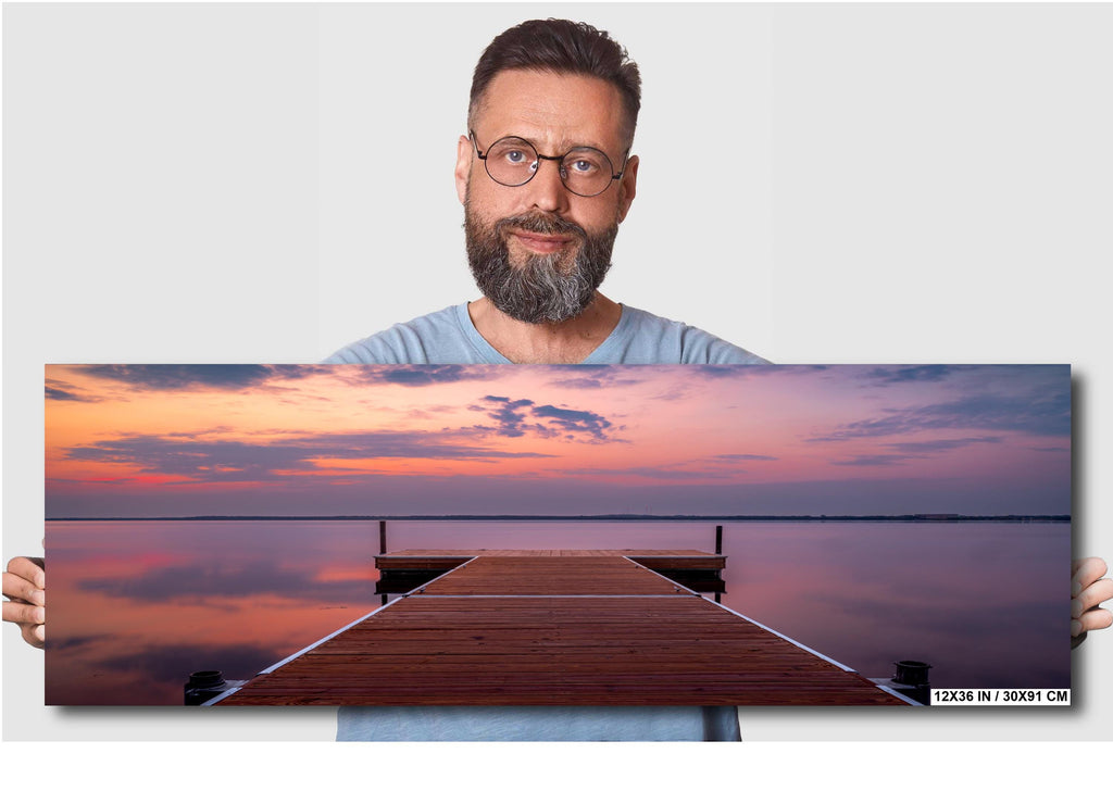 A man holding a 12x36 inches wall art of sunset over calm Lake Bemidji with dock view and pastel skies before a summer storm.