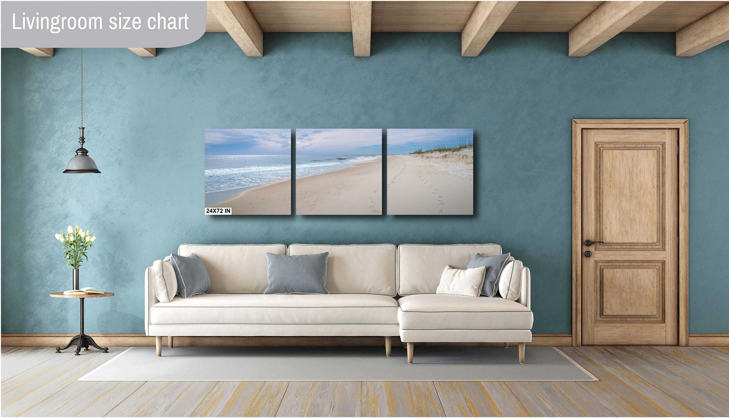 A three-panel triptych landscape photograph of a serene beach at 24x72 inches, mounted on a teal blue wall above a modern cream-colored sectional sofa in a stylish living room with wooden ceiling beams.