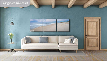 A three-panel triptych landscape photograph of a serene beach at 24x72 inches, mounted on a teal blue wall above a modern cream-colored sectional sofa in a stylish living room with wooden ceiling beams.