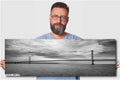 A man holding a 12x36 inch (30 x 91 cm) panoramic black and white photograph of a suspension bridge over water against a light grey background.