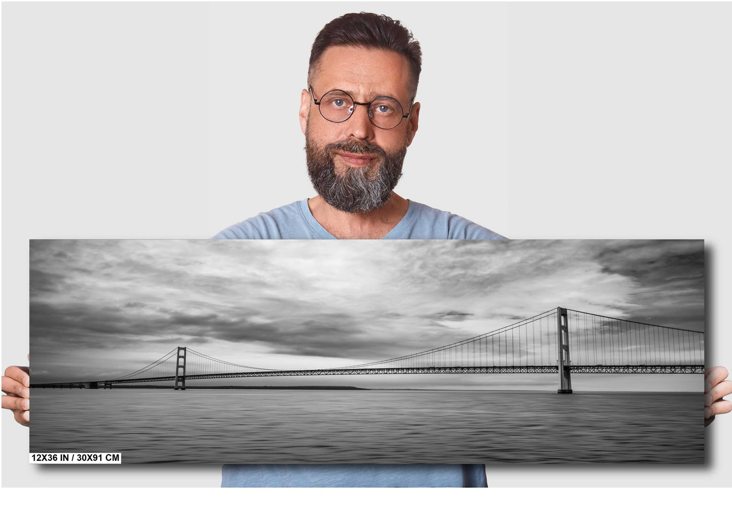 A man holding a 12x36 inch (30 x 91 cm) panoramic black and white photograph of a suspension bridge over water against a light grey background.