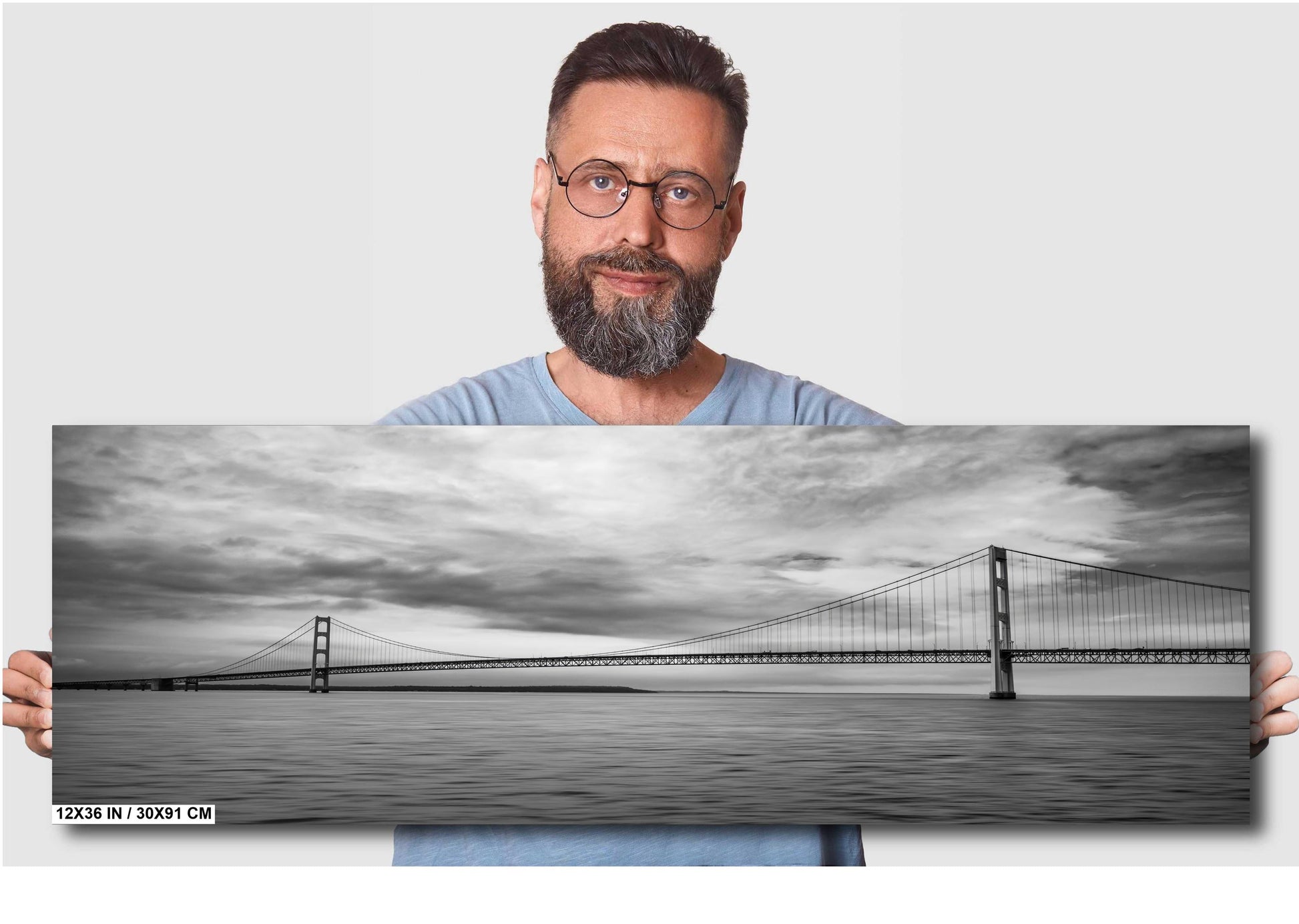 A man holding a 12x36 inch (30 x 91 cm) panoramic black and white photograph of a suspension bridge over water against a light grey background.