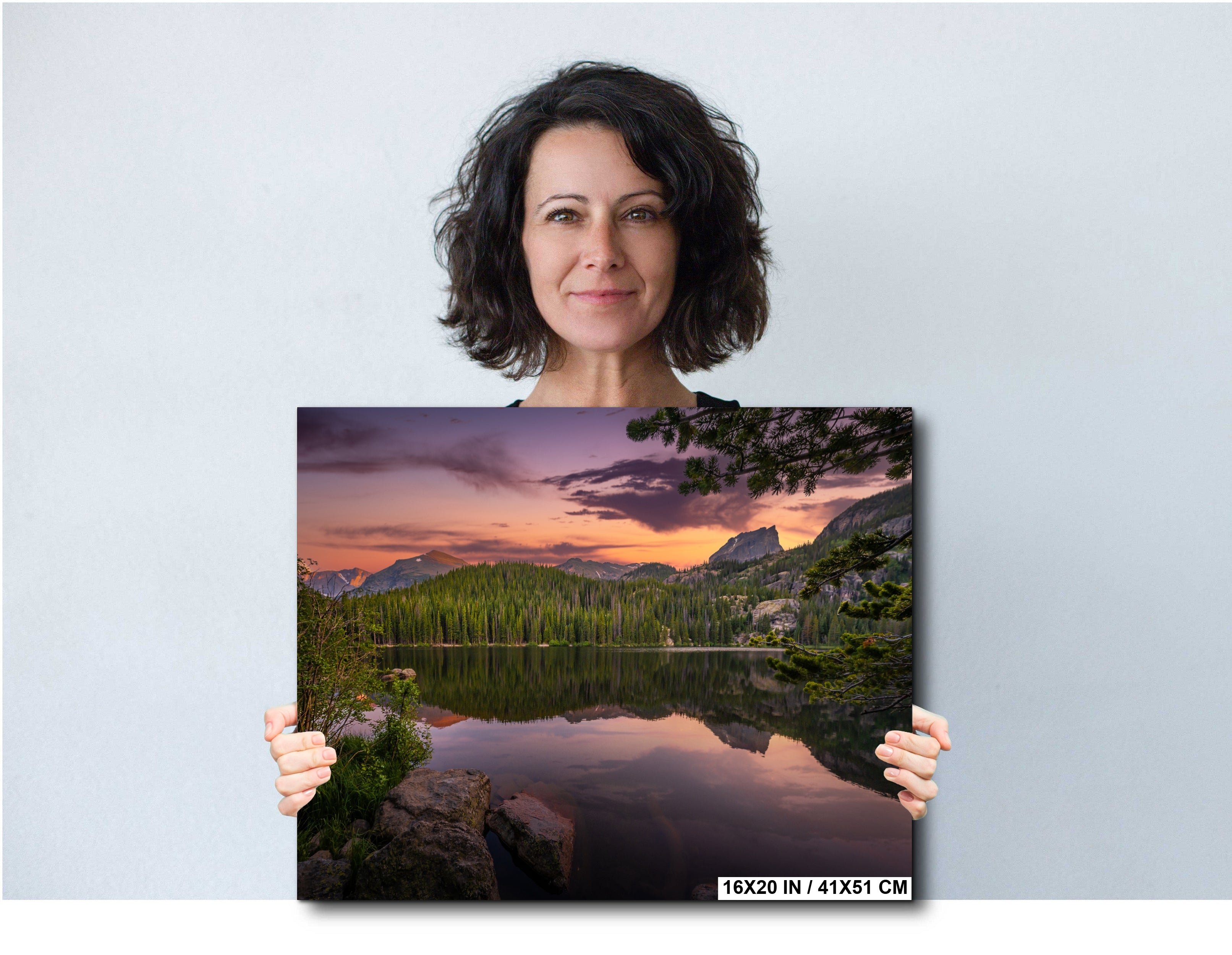 A woman holding a framed print of a serene landscape with a lake, mountains, and trees.