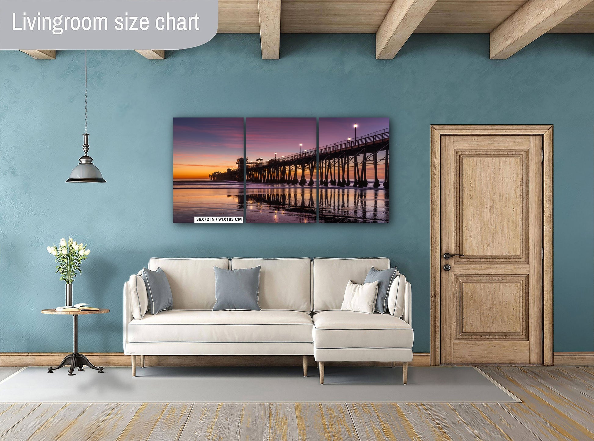 a living room with a large wall art piece featuring a serene sunset over a pier. The room has a white sofa, a wooden door, and a wooden table with a vase of flowers.