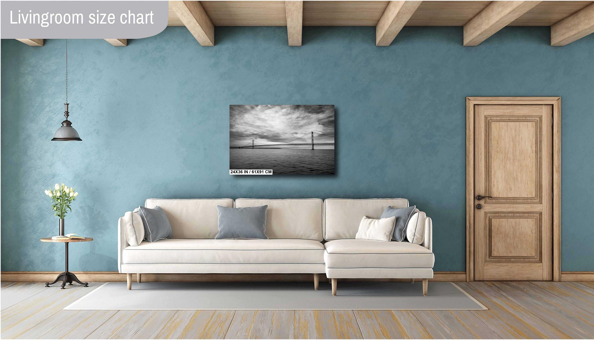 A 24x36 inch black and white horizontal art print of a suspension bridge over water, mounted on a teal blue wall above a modern cream-colored sectional sofa in a stylish living room with wooden ceiling beams.