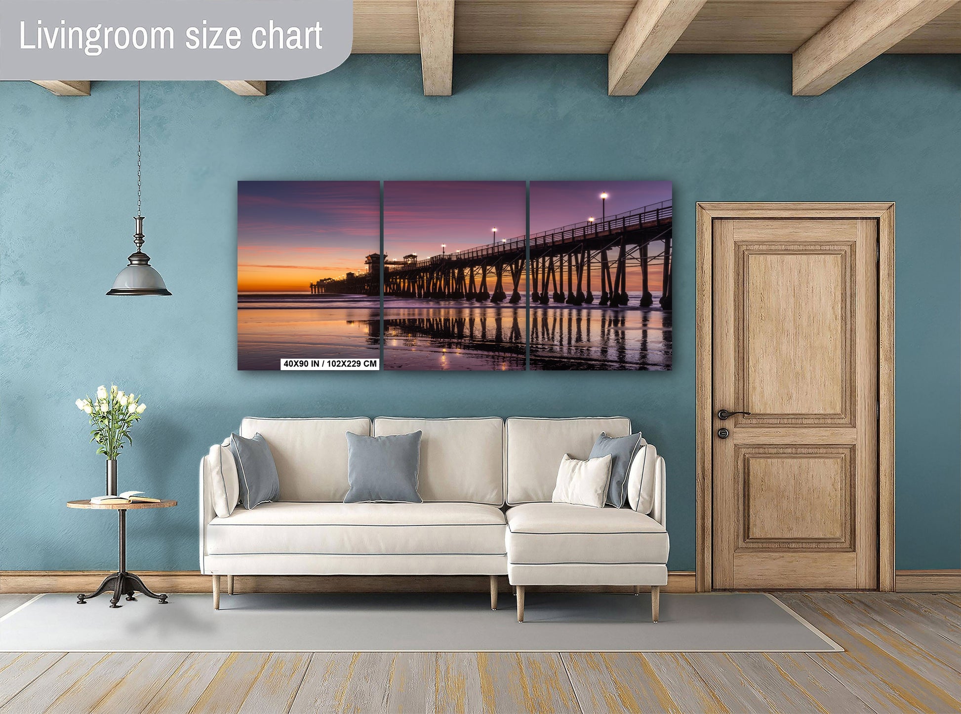 a living room with a large, colorful painting of a pier and sunset on the wall. The painting is a triptych, with the top and bottom panels showing the pier and the sky, respectively. The room has a white sofa, a wooden door, and a small table with a vase of flowers. The overall color scheme is a mix of blue and orange, creating a warm and inviting atmosphere.