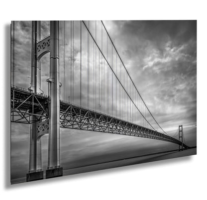 A black and white photograph of the Mackinac Bridge, capturing the engineering and grandeur of the structure with a backdrop of overcast sky and a body of water.