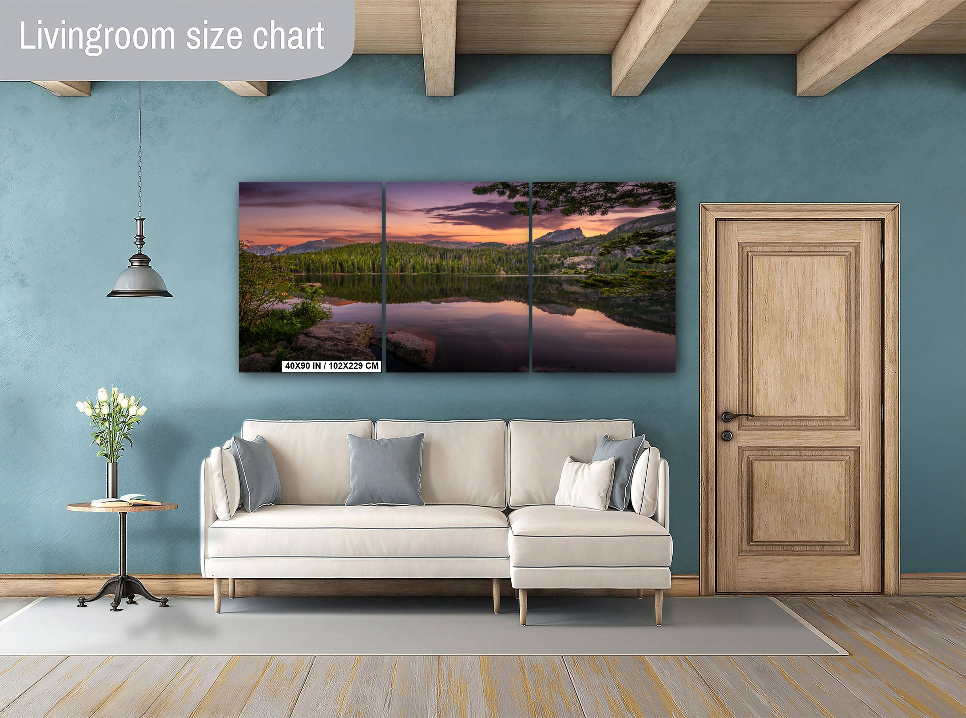 a living room with a large, three-panel landscape painting hanging on the wall. The painting features a serene lake surrounded by mountains and trees, creating a peaceful and natural atmosphere. The room is furnished with a white sofa and a wooden door, and there is a small table with a vase of flowers nearby.