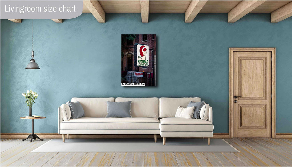 A 24x36 inch vertical art print of "The Redhead Piano Bar" neon sign mounted on a teal blue wall above a modern cream-colored sectional sofa in a stylish living room with wooden ceiling beams.