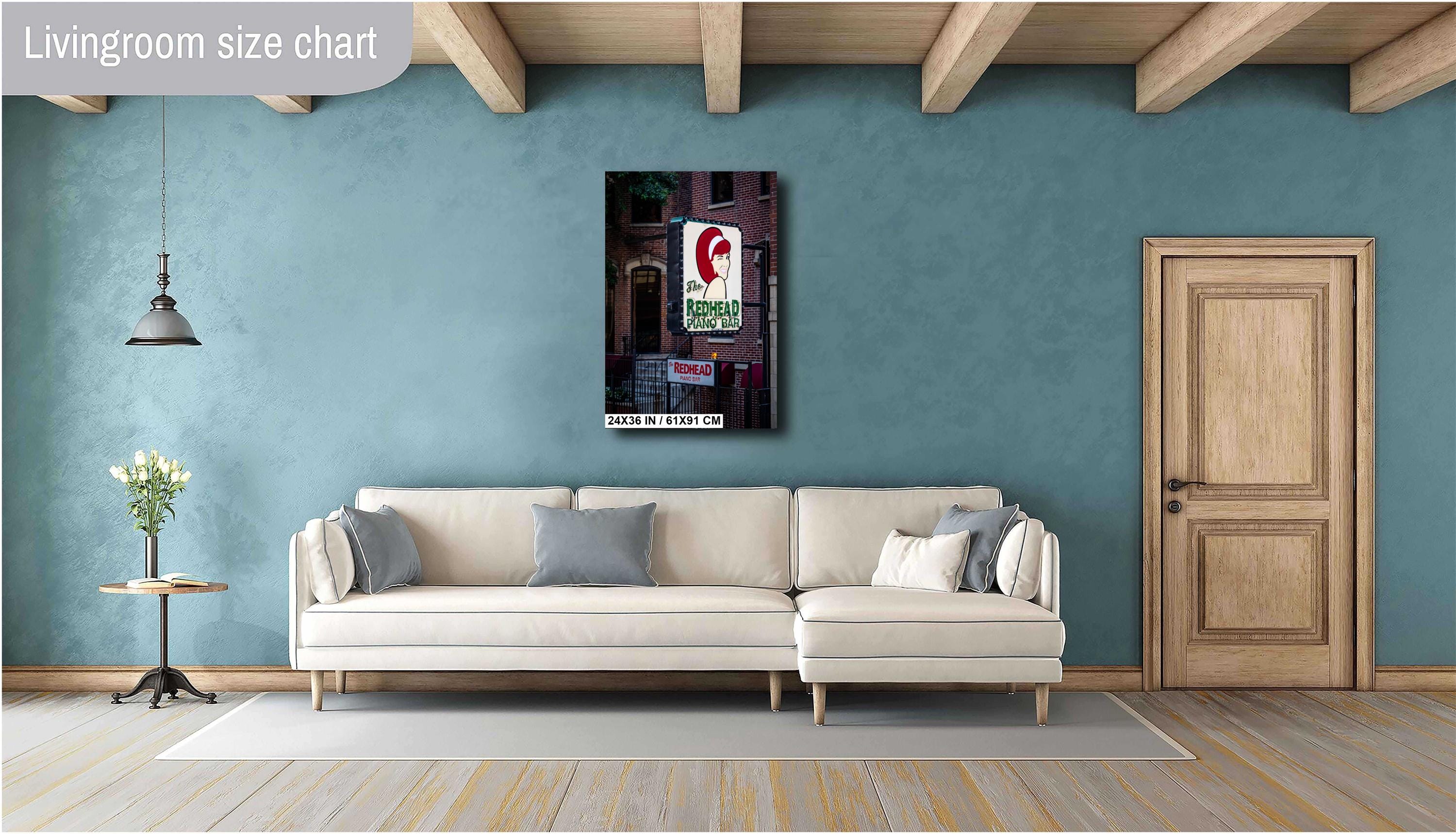 A 24x36 inch vertical art print of "The Redhead Piano Bar" neon sign mounted on a teal blue wall above a modern cream-colored sectional sofa in a stylish living room with wooden ceiling beams.