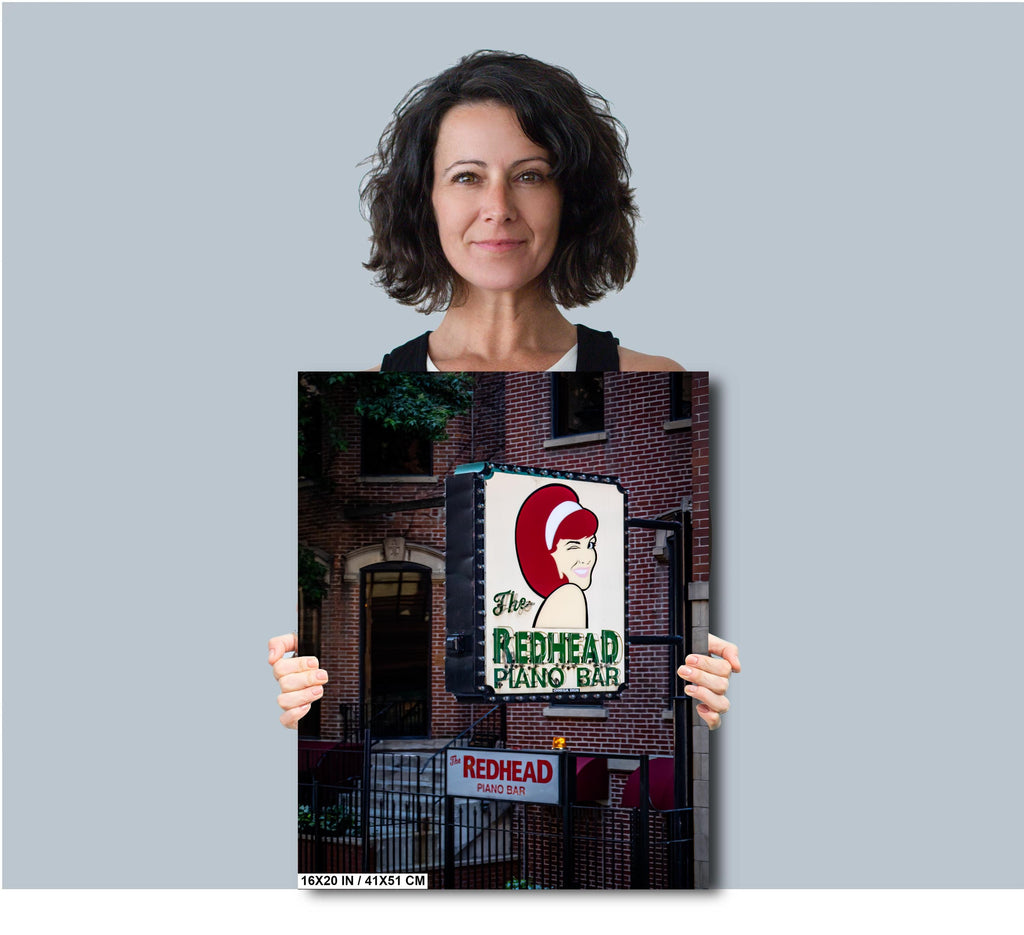 A woman with short, dark curly hair holding a 16x20 inch portrait-oriented photo print of "The Redhead Piano Bar" neon sign against a solid light grey background.