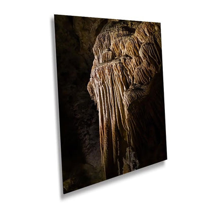 A vertical art print of a golden-brown limestone stalactite formation in a dark cave, displayed as a metal or acrylic mockup against a white background.