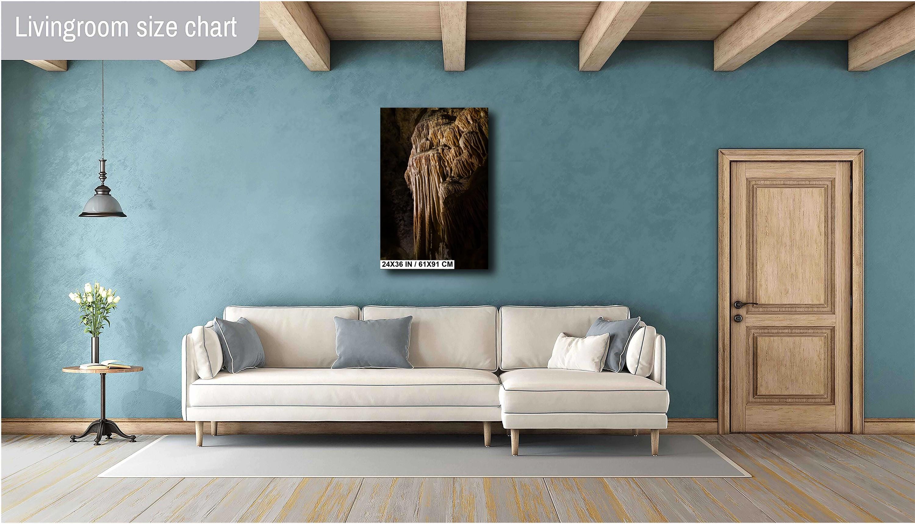 A 24x36 inch portrait-oriented art print of a golden-brown limestone stalactite formation, mounted on a teal blue wall above a modern cream-colored sectional sofa in a stylish living room with wooden ceiling beams.