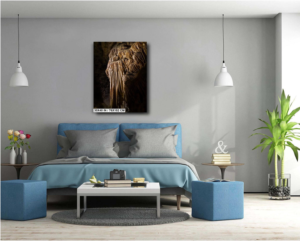 A 30x40 inch vertical art print of a golden-brown limestone stalactite formation, mounted on a light grey wall above a modern blue bed in a bright, minimalist bedroom.