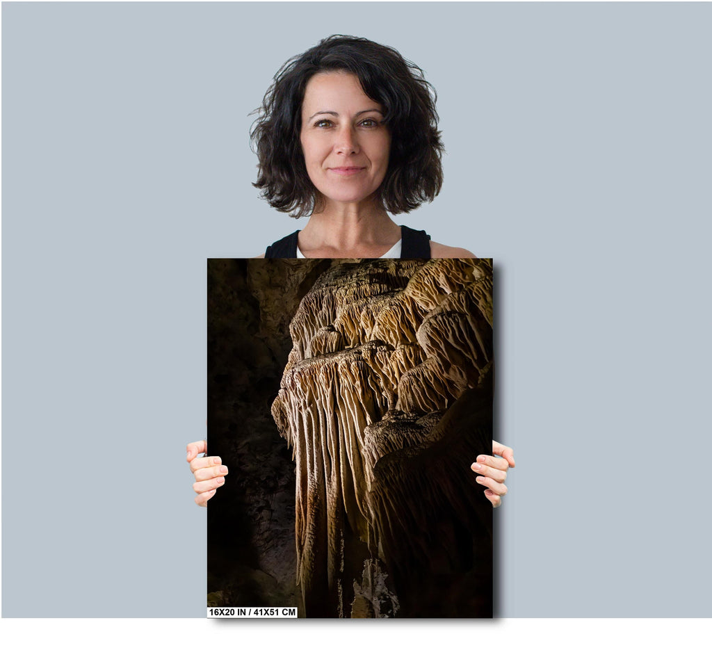A woman holding a 16x20 inch (41 x 51 cm) portrait-oriented art print of a golden-brown limestone stalactite formation against a solid light grey background.