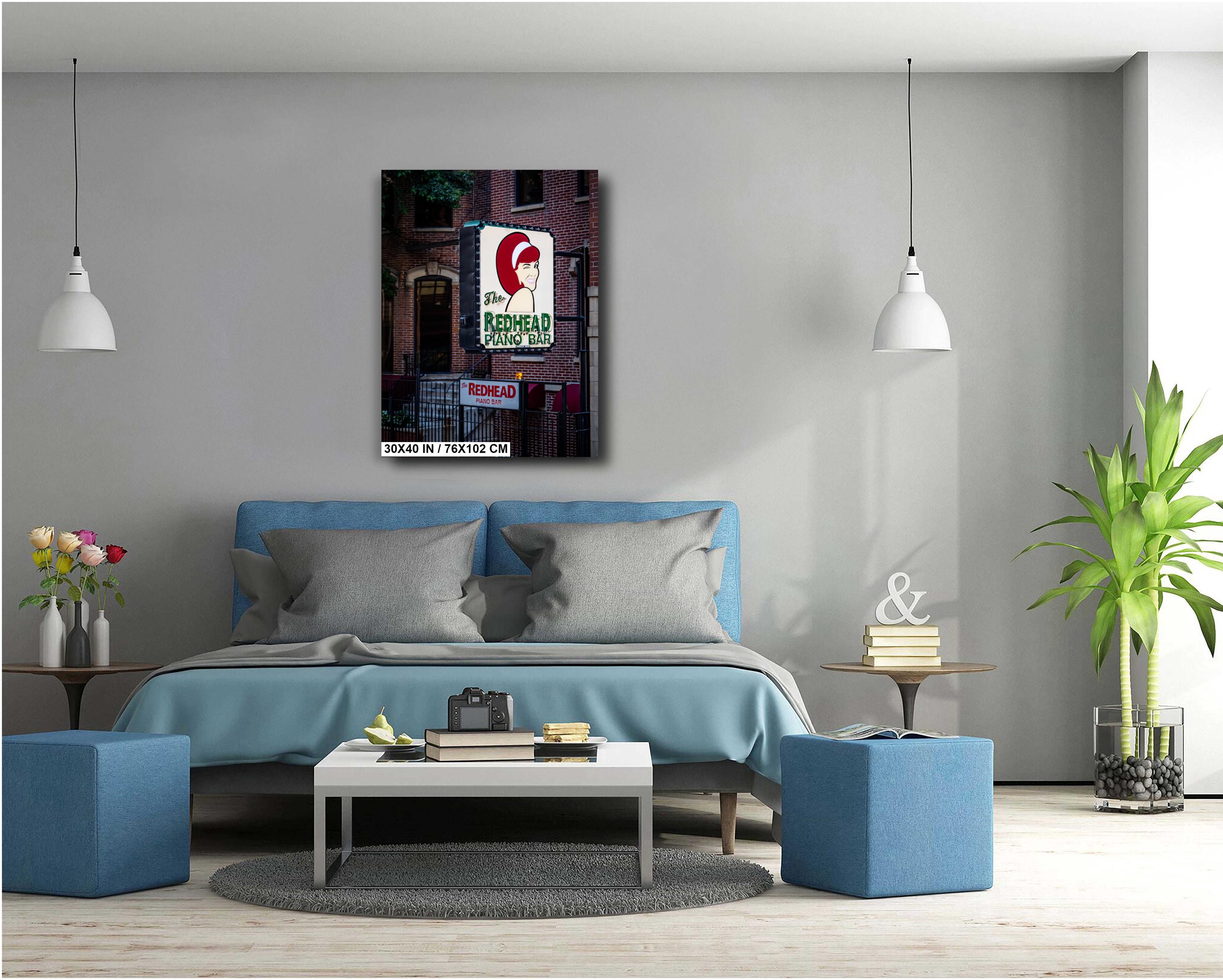 A 30x40 inch portrait-oriented art print of "The Redhead Piano Bar" neon sign mounted on a light grey wall above a modern blue upholstered bed in a minimalist bedroom.
