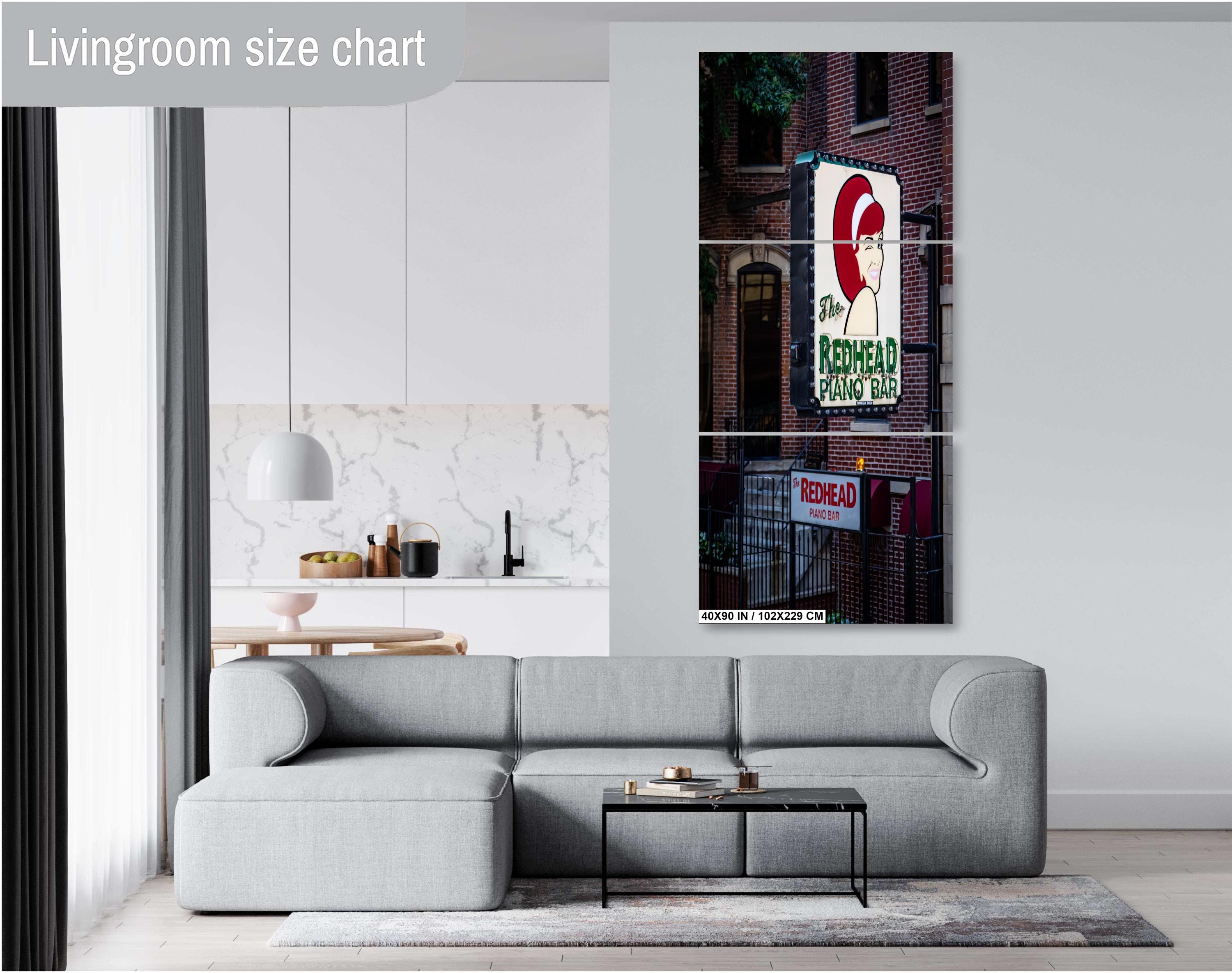 A three-panel vertical triptych art print of "The Redhead Piano Bar" neon sign, measuring 40x90 inches (102 x 229 cm), mounted on a light grey wall above a modern grey sectional sofa in a bright living room with a view of a kitchen.