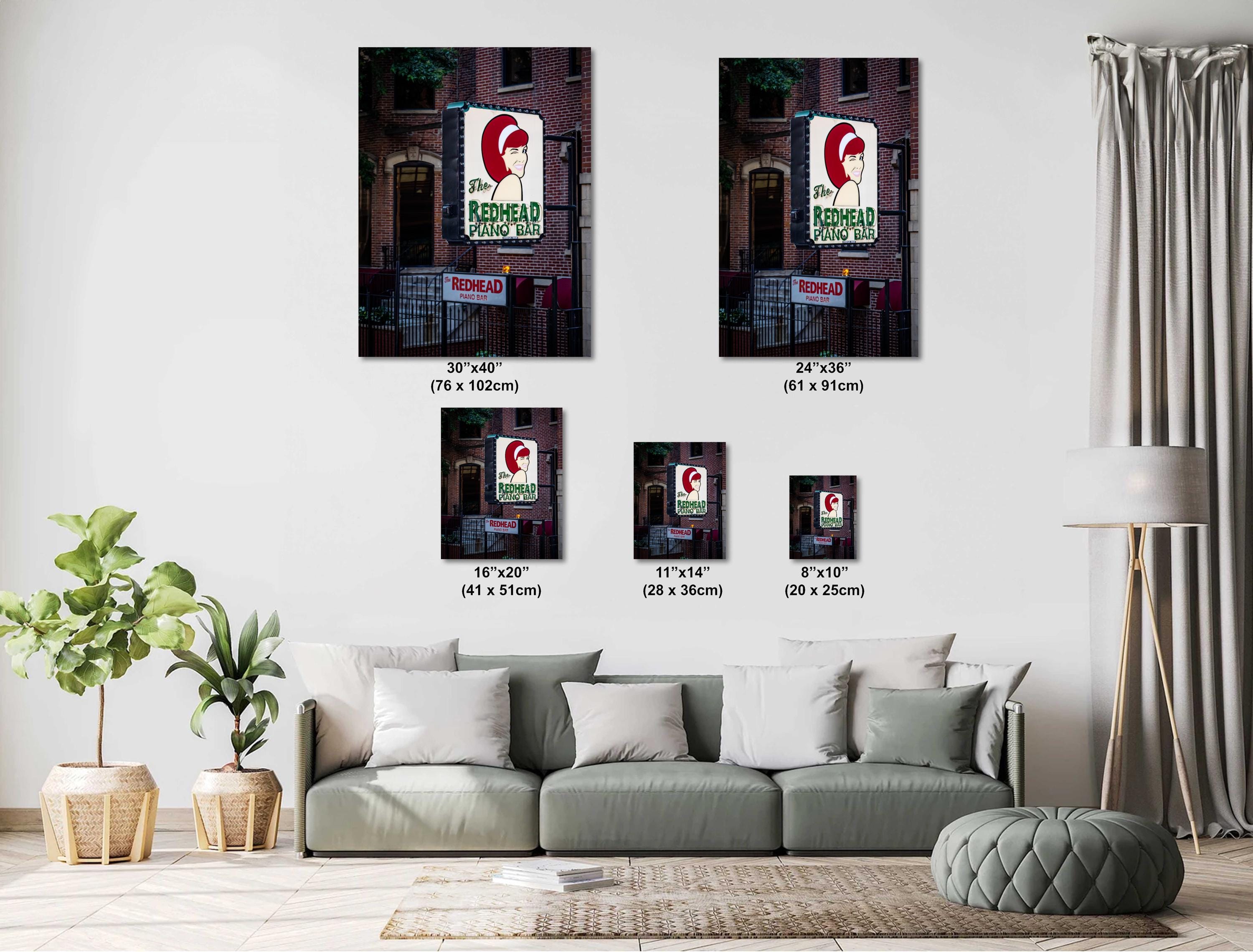 A size comparison chart featuring five different portrait-oriented prints of "The Redhead Piano Bar" neon sign, ranging from 8x10 inches to 30x40 inches, displayed on a white wall above a modern grey sectional sofa with a potted plant and floor lamp.