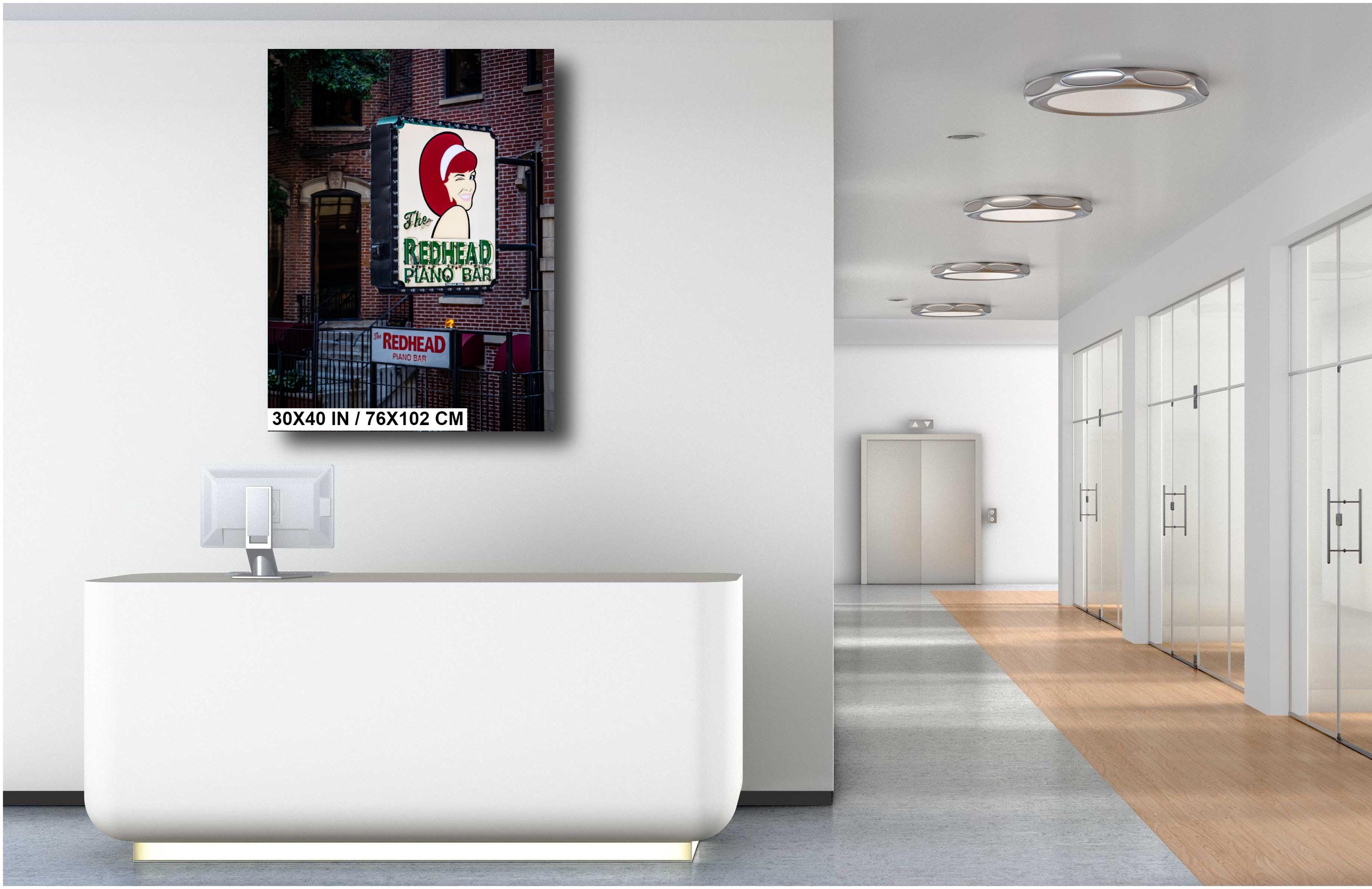 A 30x40 inch portrait-oriented art print of "The Redhead Piano Bar" neon sign mounted on a white wall above a modern white reception desk in a bright, minimalist office hallway.