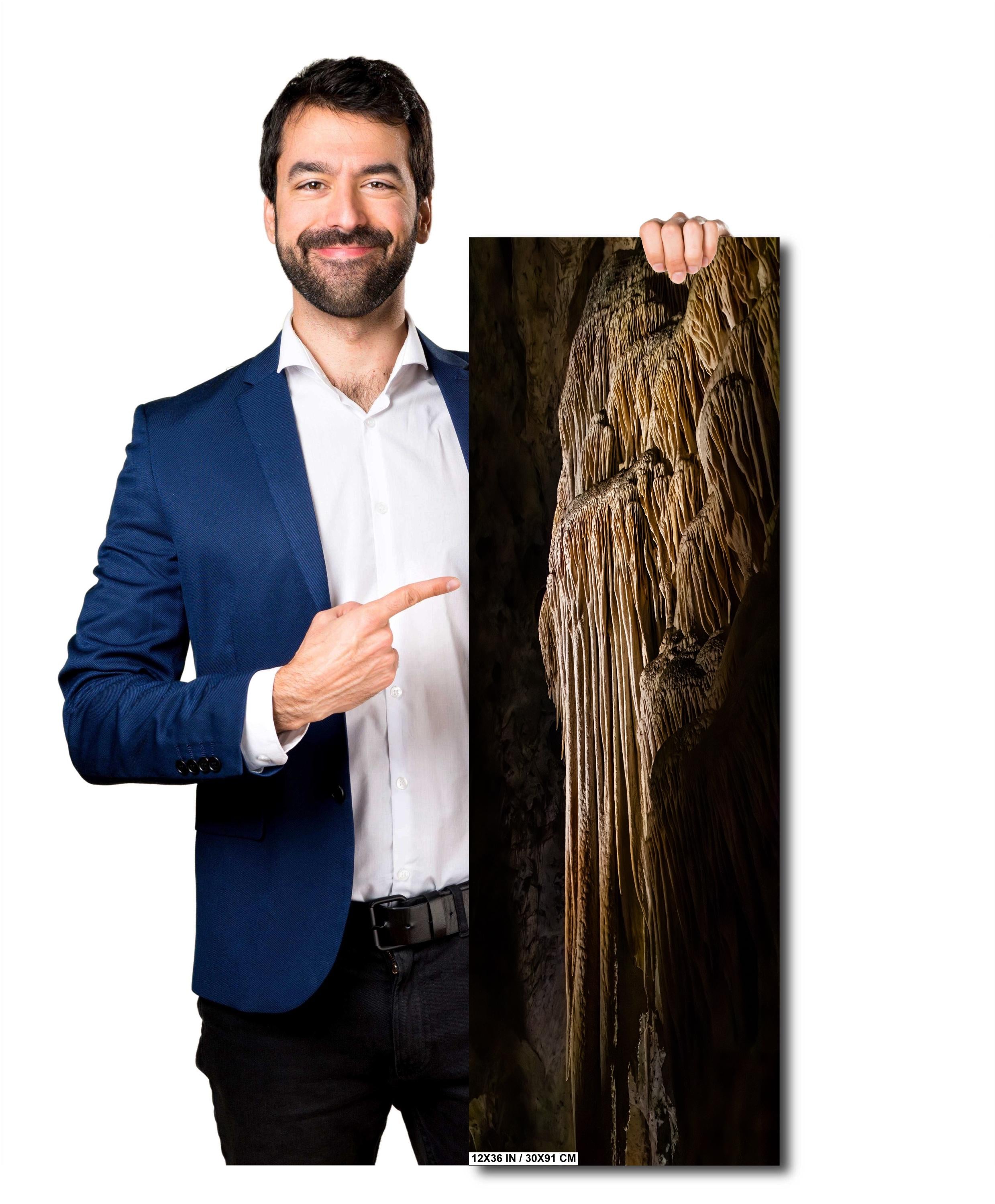 A man holding a 12x36 inch portrait-oriented art print of a golden-brown limestone stalactite formation against a solid light grey background.