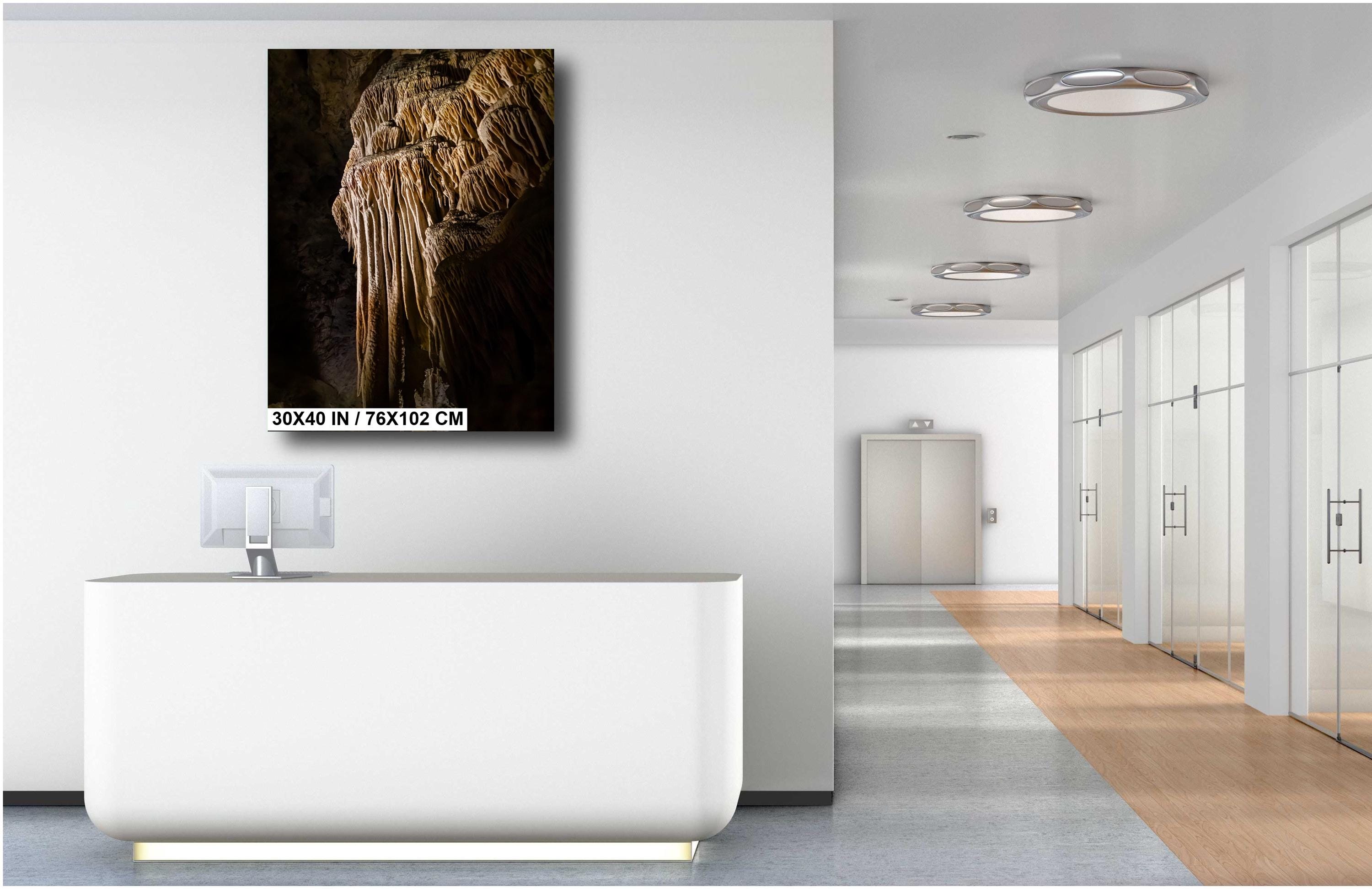 A 30x40 inch (76×102 cm) vertical art print of a golden-brown limestone stalactite formation, mounted on a white wall above a modern white reception desk in a bright, minimalist office hallway.