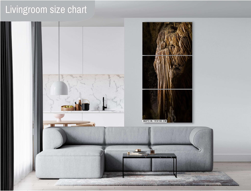 A three-panel vertical triptych art print of a golden-brown limestone stalactite formation, measuring 36x72inches (91×72 cm), mounted on a light grey wall above a modern grey sectional sofa in a bright living room with a view of a kitchen.