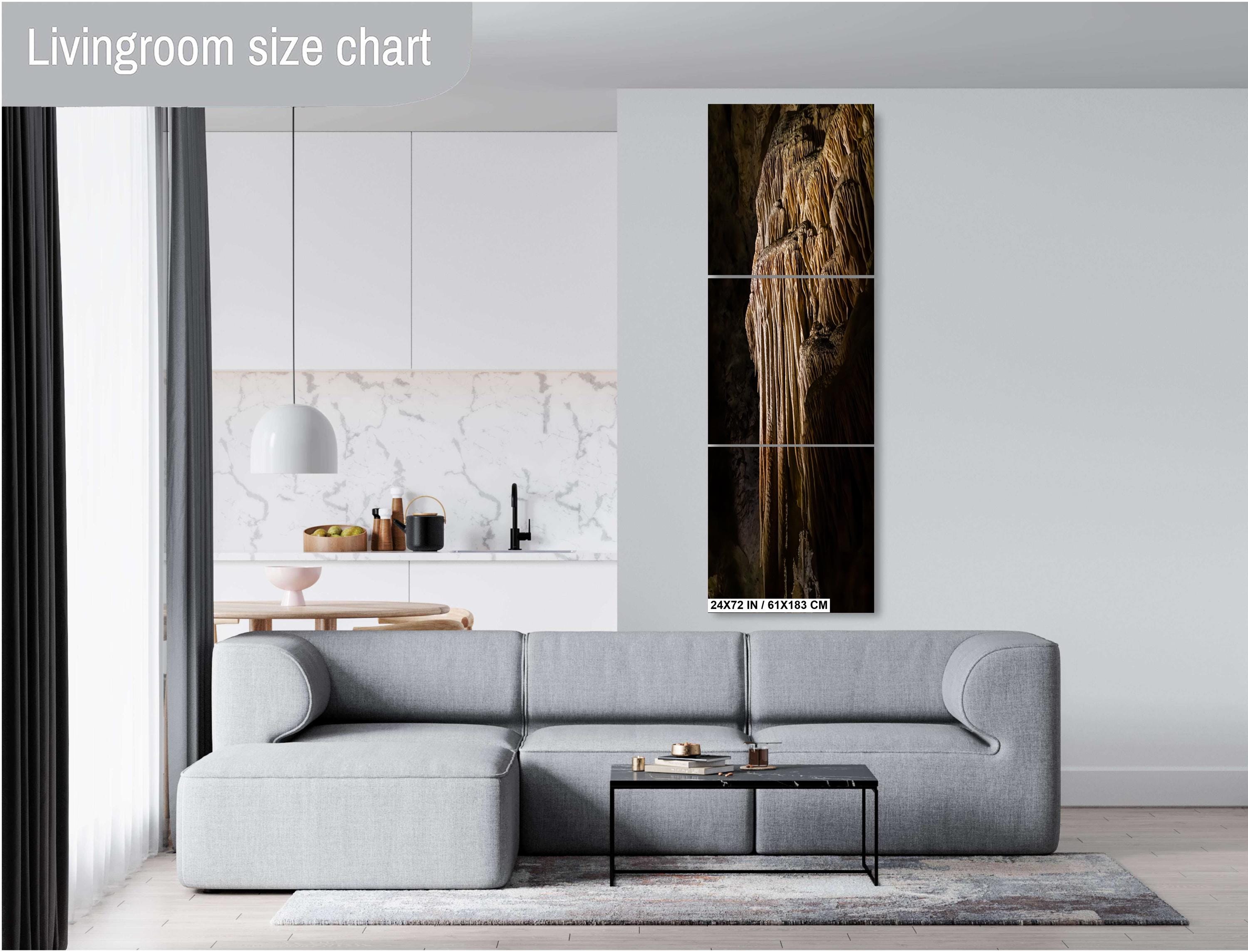 A three-panel vertical triptych art print of a golden-brown limestone stalactite formation, measuring 24x72 inches (61×183 cm), mounted on a light grey wall above a modern grey sectional sofa in a bright living room with a view of a kitchen.