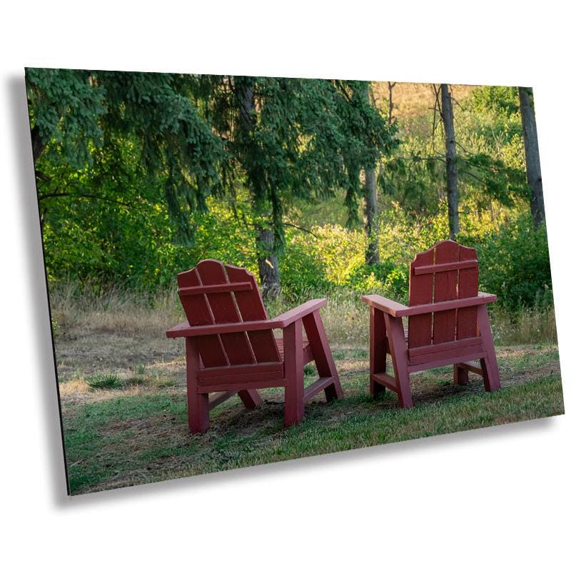 A canvas print featuring two red wooden Adirondack chairs in a natural outdoor setting with green trees and a serene expression.