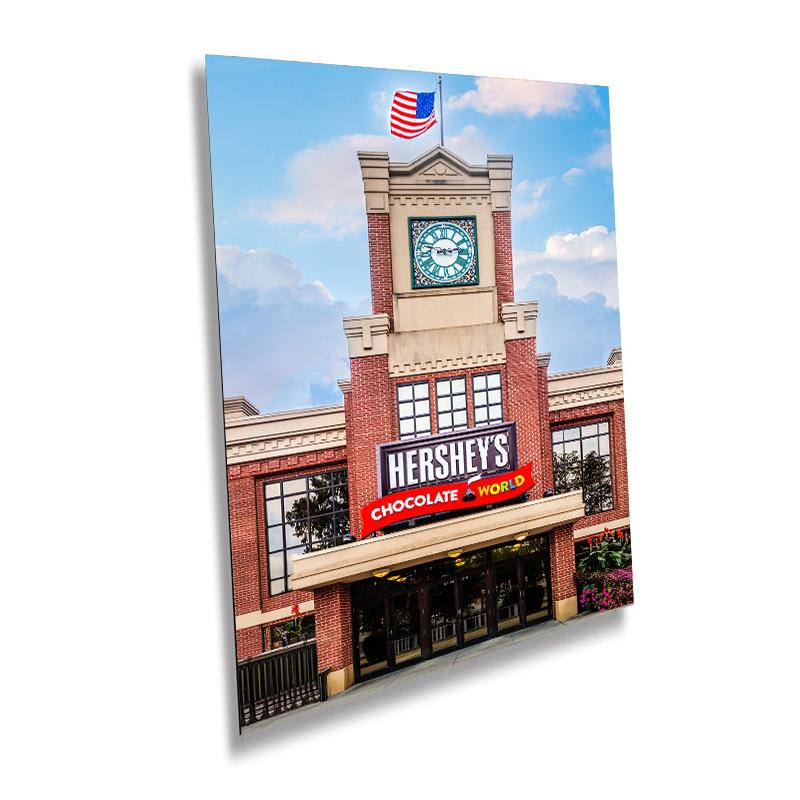 A wall art print featuring the entrance of Hershey's Chocolate World with an American flag, a clock tower, and a Hershey signboard.
